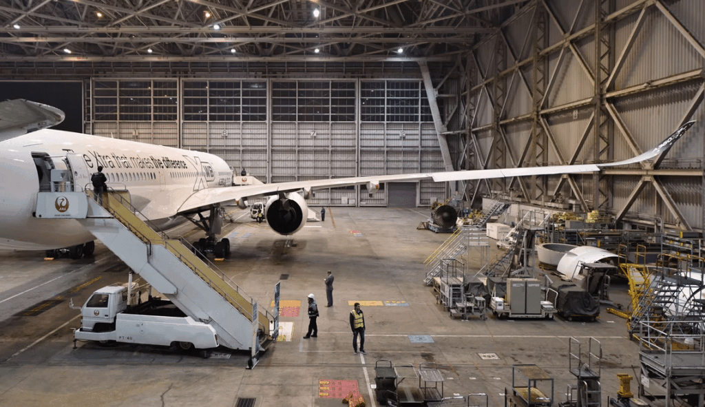 An aircraft is parked in a large hangar, with maintenance crews working. Stairs lead up to the plane, and tools are scattered around.