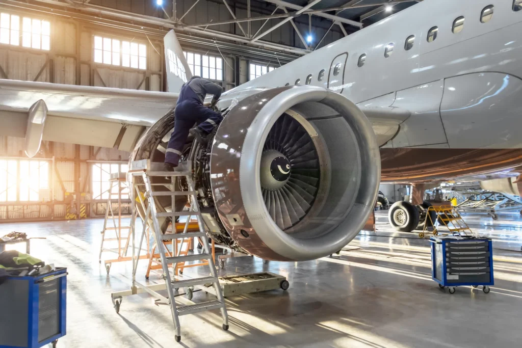 A mechanic stands on a platform working on the exposed engine of a commercial airplane inside a brightly lit hangar, with tools and equipment nearby.