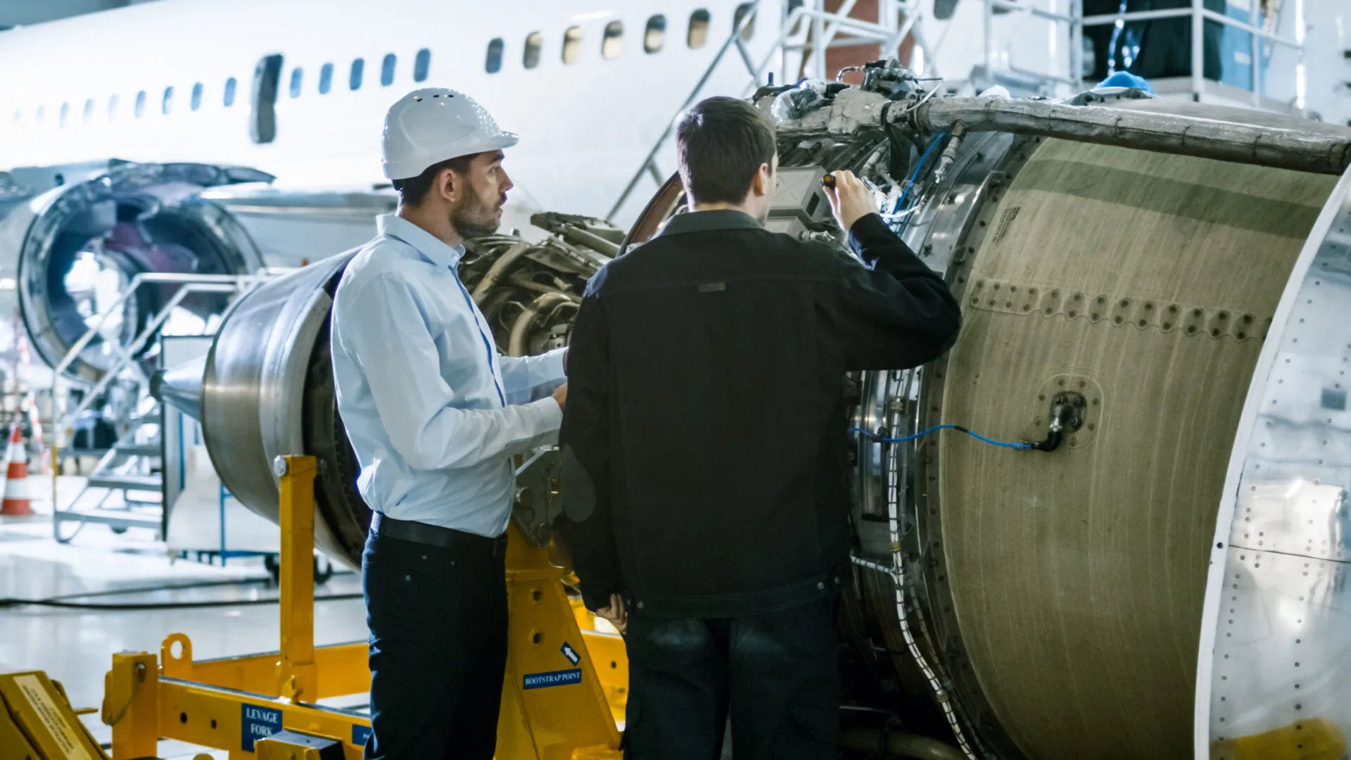 Two technicians inspect a jet engine in an aircraft maintenance hangar. One wears a hard hat and the other uses a flashlight.