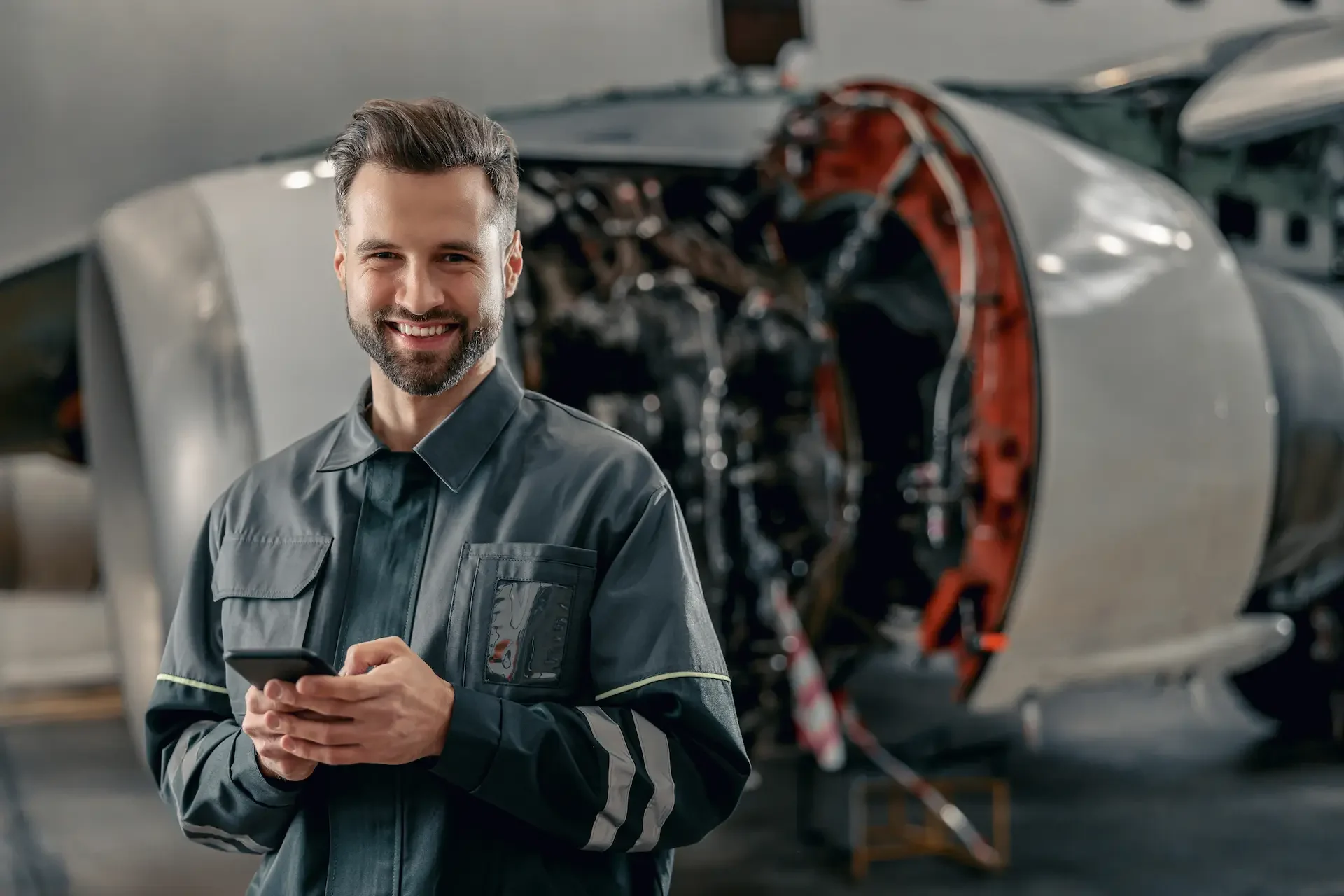 A smiling male aircraft mechanic in a dark work uniform holds a smartphone, standing in front of an airplane engine.